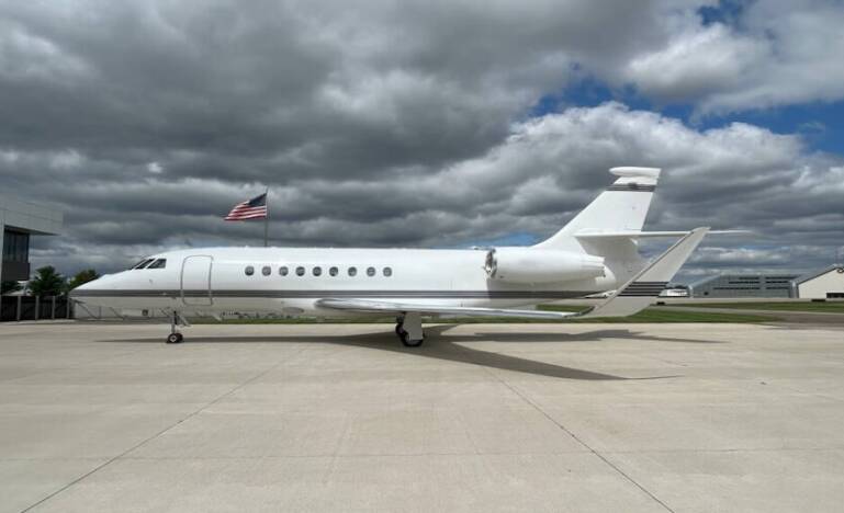 A white private jet is parked on an airport tarmac with a U.S. flag and cloudy sky in the background.