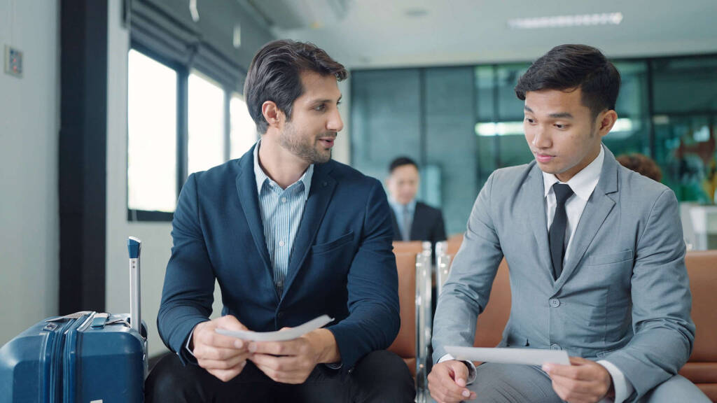 two-men-dressed-in-suits-in-the-airport-sitting-in-terminal-looking-at-paper-while-talking